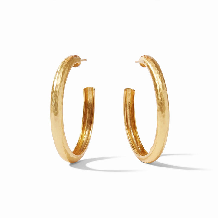 Handcrafted gold jewelry, a pair of thick hoop earrings positioned on a white background featuring a distinctive hand-hammered texture, high-shine polished finish, and a bold tubular C-shape silhouette