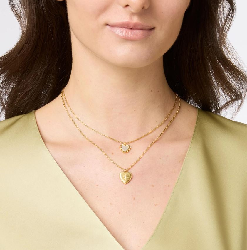 Studio photograph of a woman wearing two layered gold heart necklaces on her neck featuring a small scalloped heart with a white center and a larger gold heart with a starburst motif against an olive green satin blouse