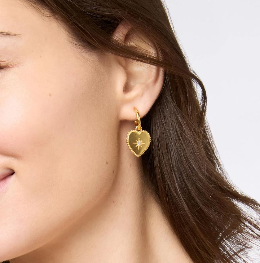 Close-up photograph of a gold heart-shaped earring worn on a woman's ear featuring a beaded edge and a central starburst motif with a sparkling white crystal