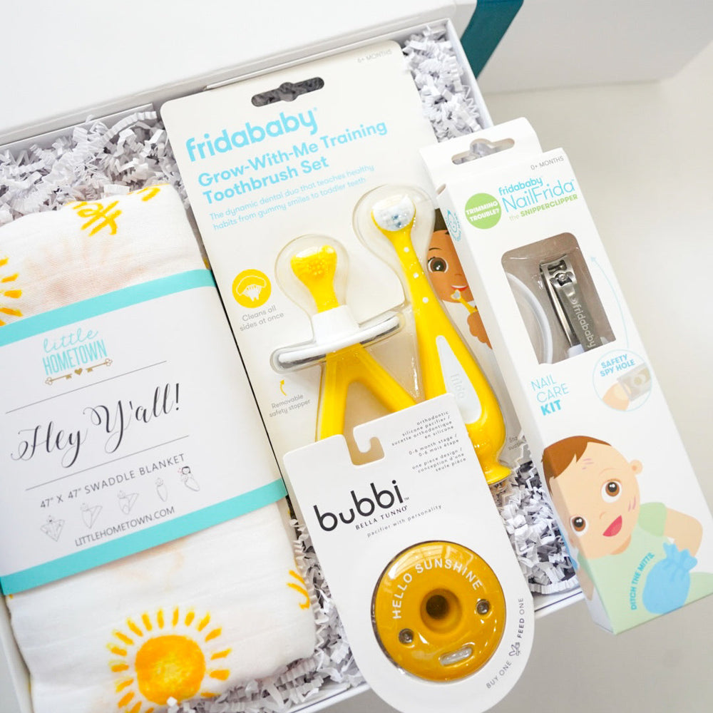 Commercial product photograph of a baby gift box featuring a Little Hometown sun-patterned swaddle, yellow Fridababy training toothbrushes, and a Bella Tunno Hello Sunshine pacifier nestled in white shredded paper filler