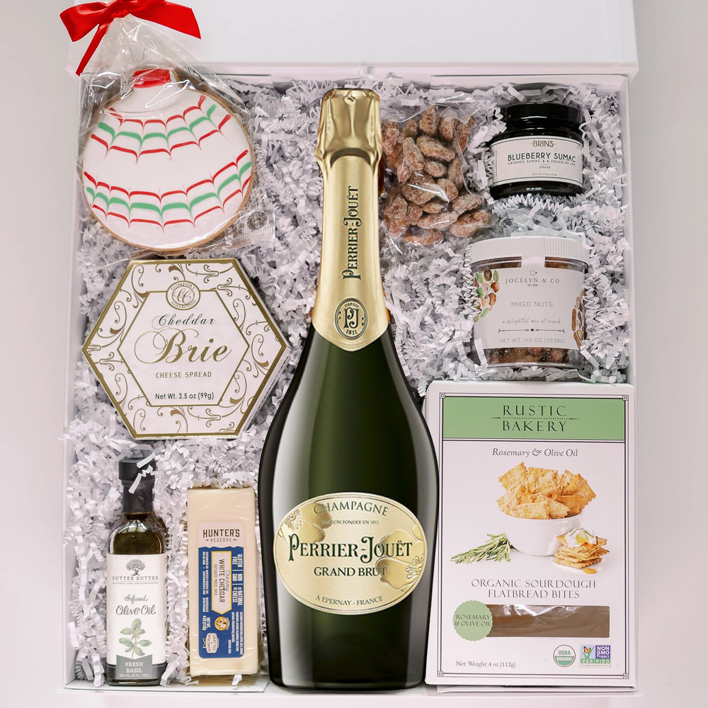 Photograph of a luxury gourmet gift basket in a white box featuring a bottle of Perrier-Jouët Grand Brut Champagne, a festive red and green scalloped iced cookie, and artisanal snacks like Rustic Bakery flatbread bites nestled in white crinkle paper