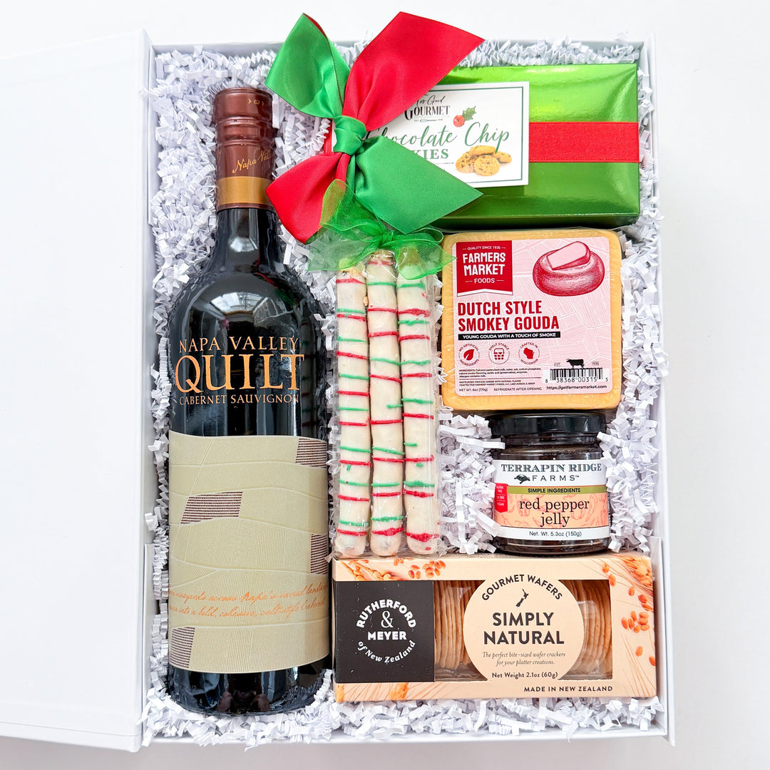 A curated gift box filled with assorted treats and snacks, including a bottle of red wine, candy canes, a green gift box tied with a red bow, a jar of spread, and packaged snacks, set against a plain white background.