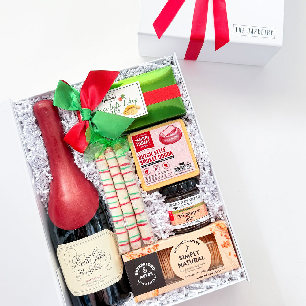 A curated gift box set from The Gathering, featuring a bottle of sparkling red wine, a bottle of peppermint candy, artisanal cheeses, crackers, and chocolates, arranged in white crinkle paper inside an open white gift box with a red ribbon. A matching closed white gift box with a red ribbon bow is shown in the background on a clean white surface.