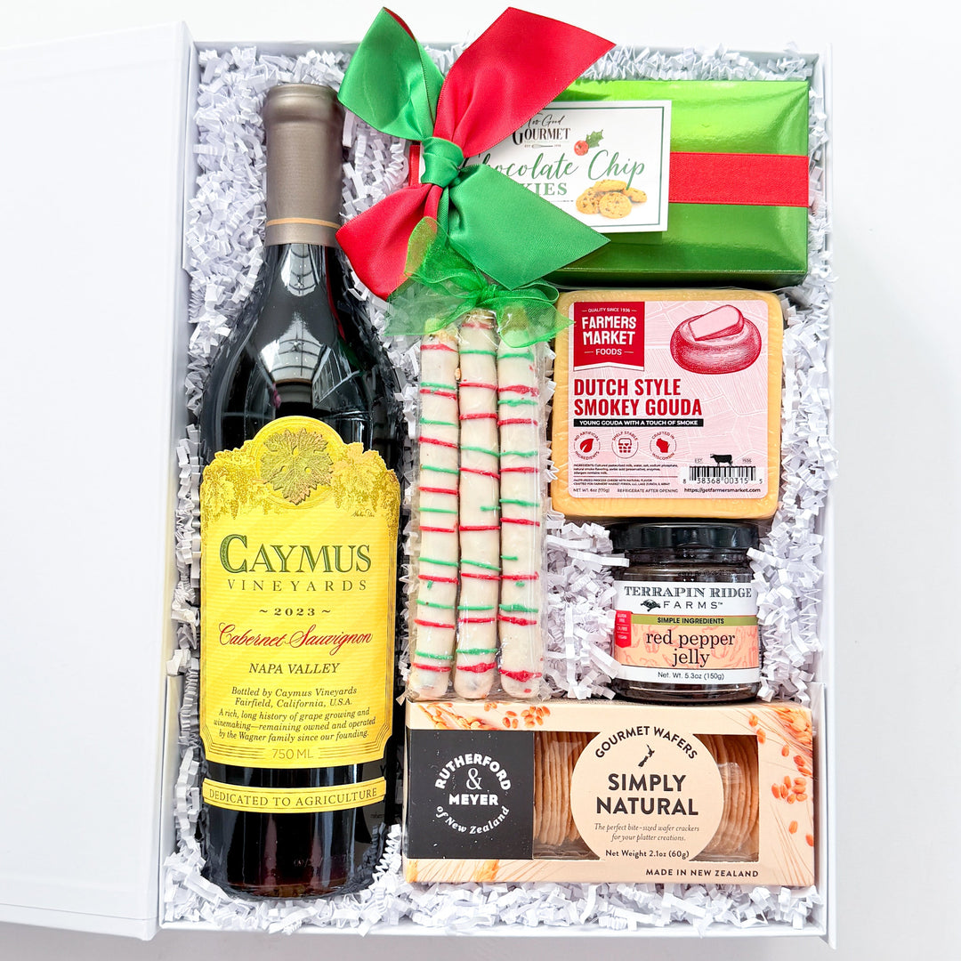 An open gift box filled with assorted holiday treats and a bottle of wine, arranged on a white background. Visible items include a bottle of Caymus Red Blend Sauvignon, wrapped chocolates, candy canes, a green gift box tied with a red ribbon, a jar of peppermint, and packaged snacks.