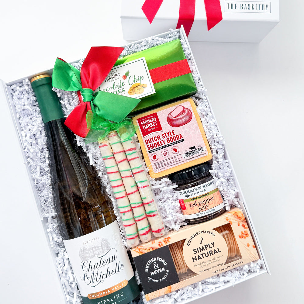 An open white gift box tied with a bright red ribbon, filled with assorted gift items including a bottle of wine, candy canes, a green and red wrapped item, and packaged snacks. A closed white box with the same red ribbon bow sits behind it on a clean white background.