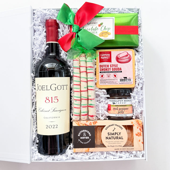 A curated gourmet gift box containing a bottle of Joel Gotti 815 California Sauvignon, Dutch Style Sandwich Cookies, a chocolate chip cookie, a jar of jam, and a Simply Natural chocolate bar, arranged in white crinkle paper with a red and green ribbon.