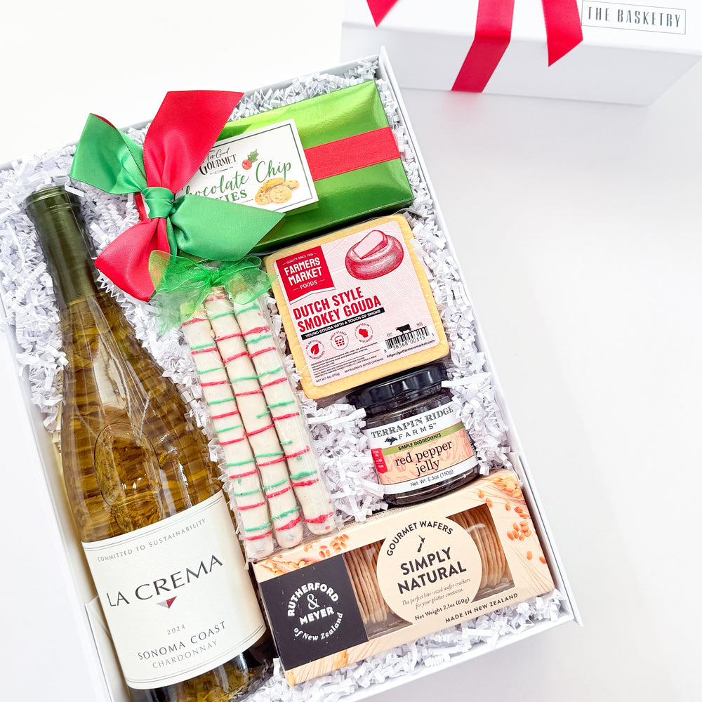 A curated gift box set on a white background, featuring a bottle of La Crema Sauvignon Blanc, a green and red wrapped item, striped candy canes, a jar of preserves, and packaged snacks in white crinkle paper. A white gift box with a red ribbon bow sits behind the open box.
