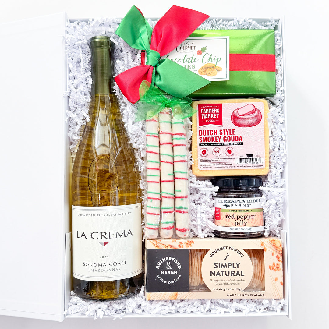 A curated gift box set containing a bottle of La Crema white wine, a green chocolate bar with a red bow, candy canes, Dutch Style Candy Cane Cookies, a jar of peppermint, and a Simply Natural soap, arranged in white crinkle paper.