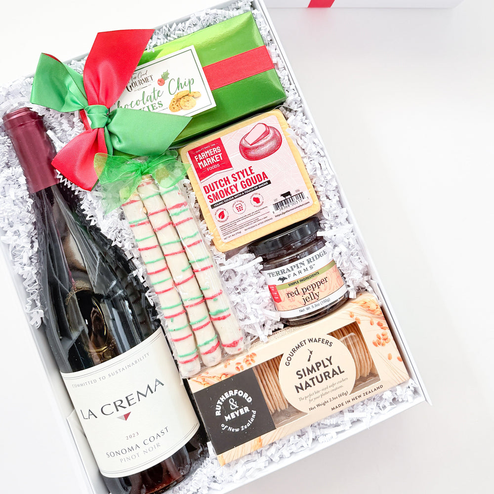 A curated gift box set from The Basaltory, featuring a bottle of La Crema wine, a jar of chocolate spread, a bag of Simply Natural cookies, candy canes, and wrapped treats, arranged in white crinkle paper inside an open white box with a red ribbon. The closed white gift box with its red ribbon is shown in the background.