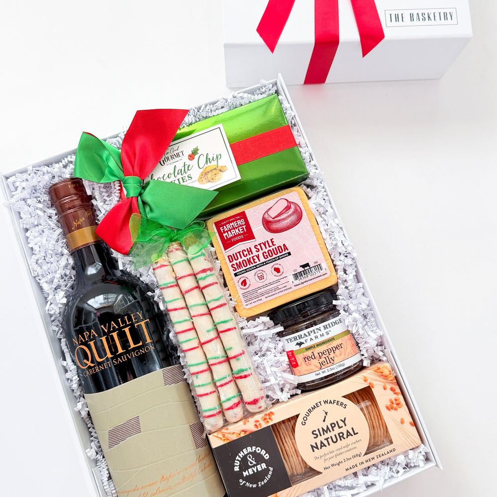 A curated gift box set from The Gift Box Store, featuring a bottle of Quillit wine, a green and red wrapped chocolate, striped candy canes, and assorted chocolates and crackers, displayed next to its white box with a red ribbon.