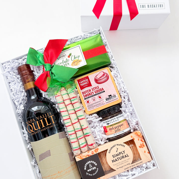 A curated gift box set from The Gift Box Store, featuring a bottle of Quillit wine, a green and red wrapped chocolate, striped candy canes, and assorted chocolates and crackers, displayed next to its white box with a red ribbon.