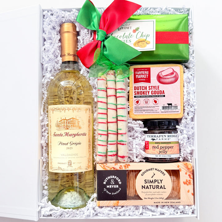 An open gift box filled with assorted gourmet items, including a bottle of Saint Marguerite Pinot Grigio white wine, a green and red wrapped item tied with a red bow, striped candy canes, a jar of Dutch Style Hammy Cocoa, a small jar of pepperoni, and packaged snacks, all arranged on white paper fill.