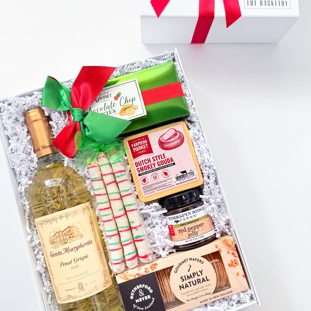 A curated gift box set on a white background, featuring a bottle of wine, candy canes, gourmet snacks, and a white gift box with a red ribbon. The wine bottle label reads 'Saint Marguerite Pinot Grigio', and the box lid has 'L'ARTISANAT' printed on it.