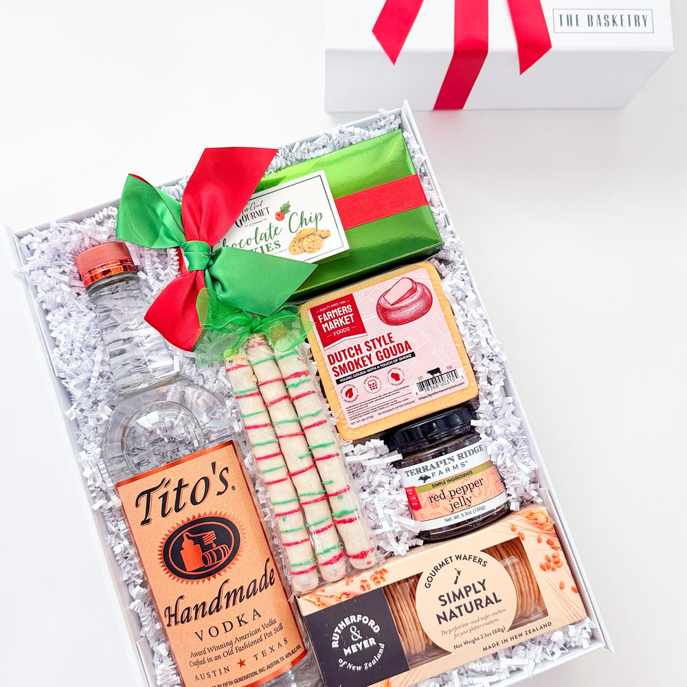 A curated gift box set on a white background, featuring a bottle of Tito's Handmade Vodka, a green and red wrapped chocolate bar, a jar of gourmet spread, candy canes, and a packaged snack, arranged in white crinkle paper next to an open white gift box with a red ribbon.