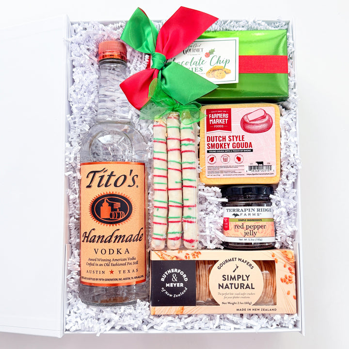 A curated gift box filled with assorted snacks and drinks, tied with a red and green bow. Visible items include a bottle of Tito&