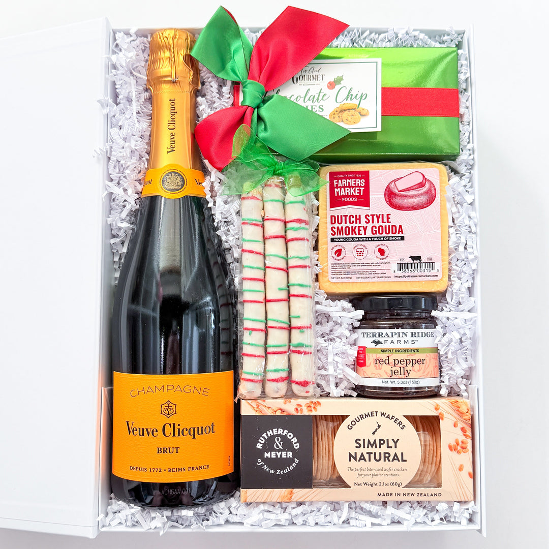 A curated gift box set containing a bottle of Vene Cliquot Brut sparkling wine, a green chocolate chip cookie, Dutch Style Smoked Gouda cheese, a jar of pepper jack, and a box of Simply Natural crackers, arranged in white crinkle paper with a red and green bow.