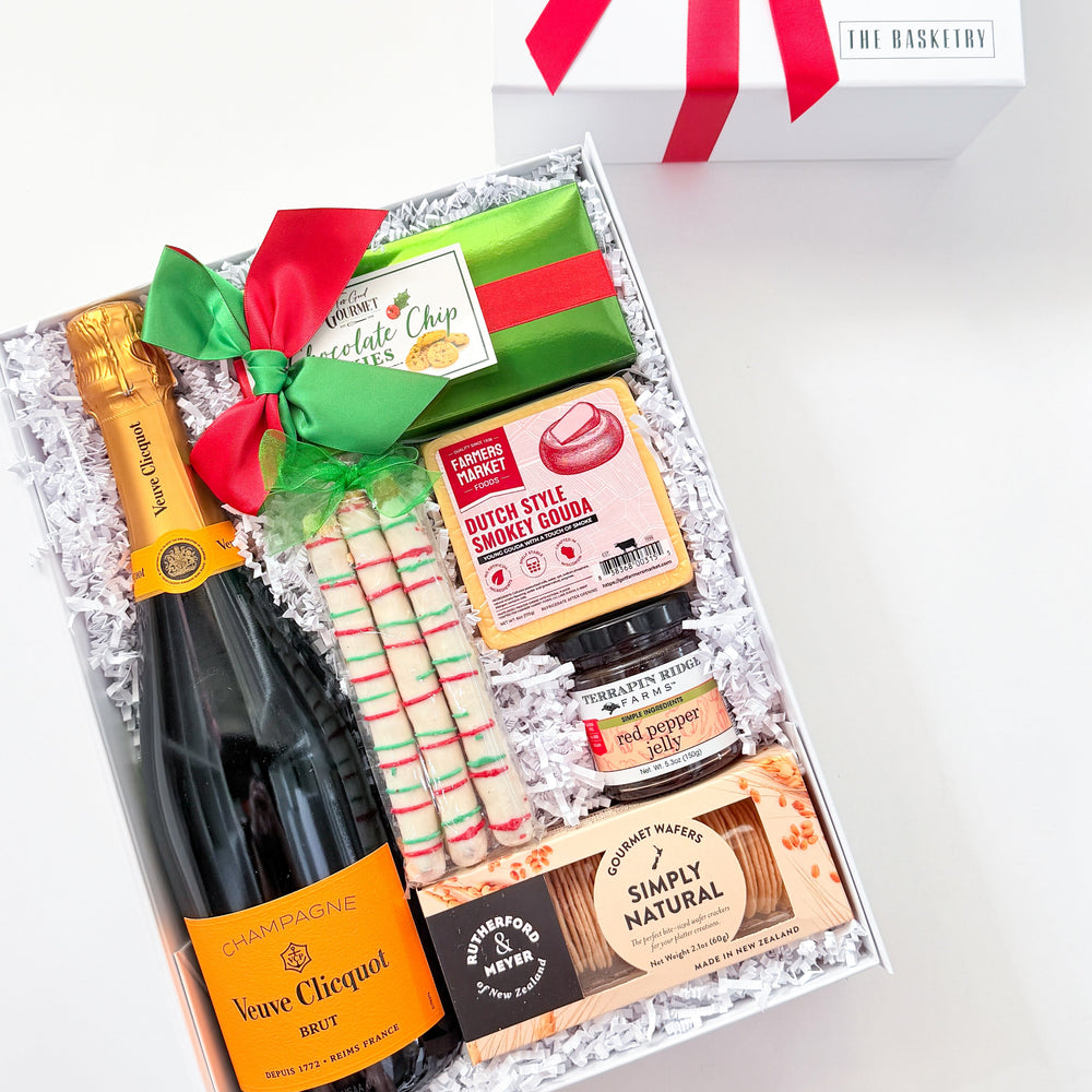 A curated gift box set on a white background, featuring an open box with a bottle of Bréme Clérical champagne, assorted chocolates, candy canes, and a red/green ribbon, alongside a closed white gift box with a red bow. The brand 'JBD GIFT SETTING' is visible on the closed box lid.