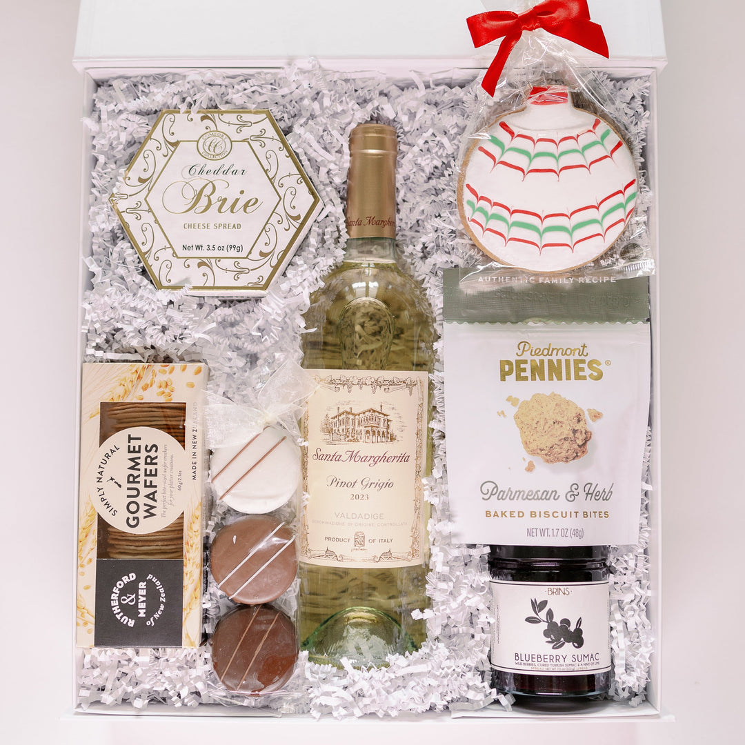 A professional studio photograph of a luxury holiday gift box featuring Santa Margherita Pinot Grigio and Piedmont Pennies biscuits alongside a red-ribboned ornament cookie with colorful scalloped icing nestled in white crinkle paper