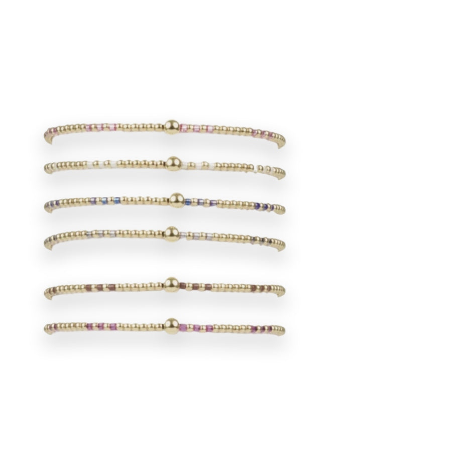 Beaded jewelry collection of six thin bracelets featuring gold spherical accents and colorful metallic beads in fuchsia and chocolate displayed on a white cylindrical stand