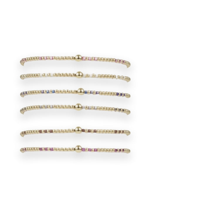Beaded jewelry collection of six thin bracelets featuring gold spherical accents and colorful metallic beads in fuchsia and chocolate displayed on a white cylindrical stand