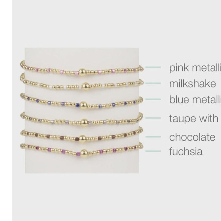 Studio product photograph of six stacked beaded bracelets displayed on a white cylindrical stand featuring central gold-tone spherical beads and colorful accents in pink metallic, chocolate, and fuchsia seed beads
