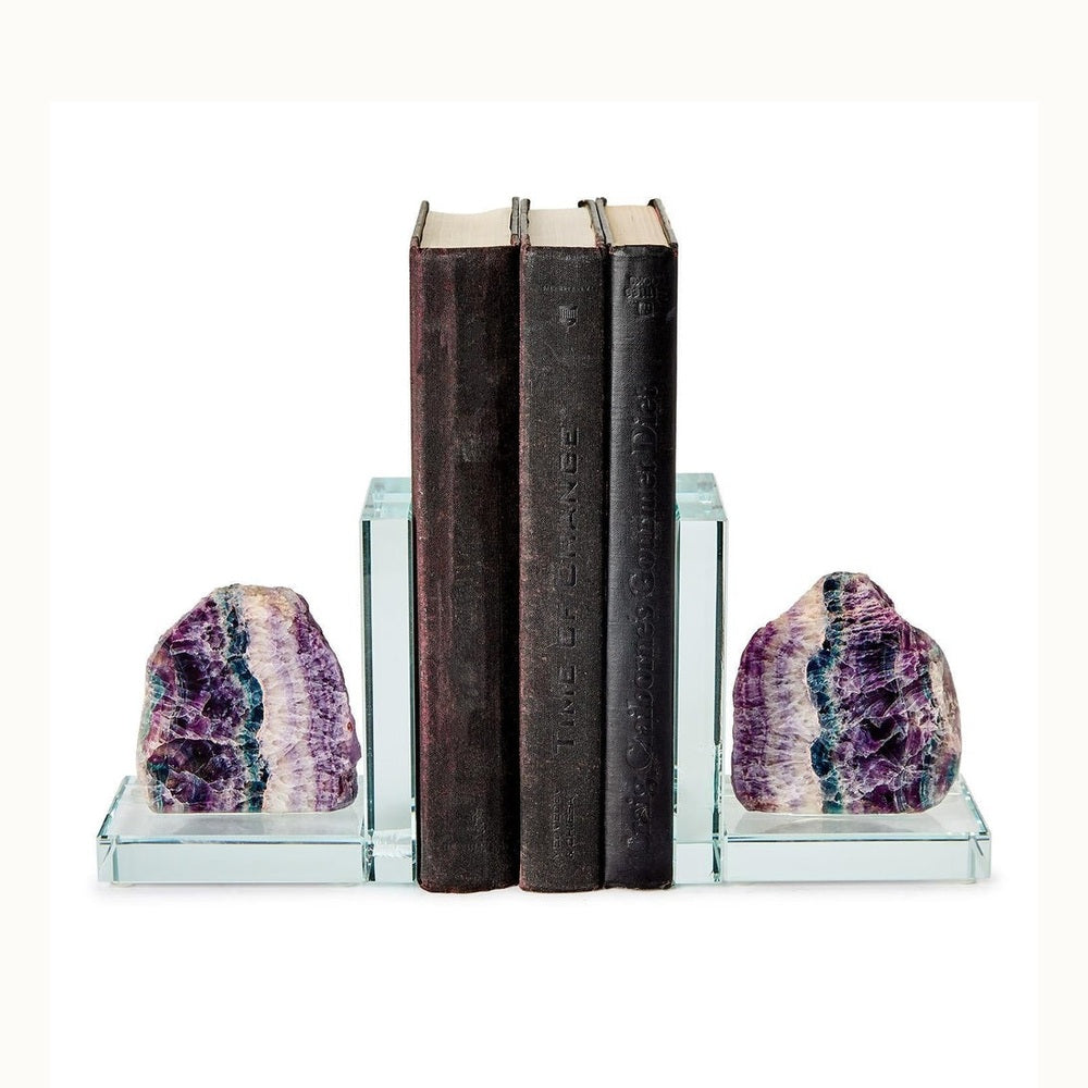 Polished glass and rainbow fluorite bookends supporting three dark vintage books against a white background featuring vibrant banded layers of purple and teal stone mounted on thick L-shaped transparent bases