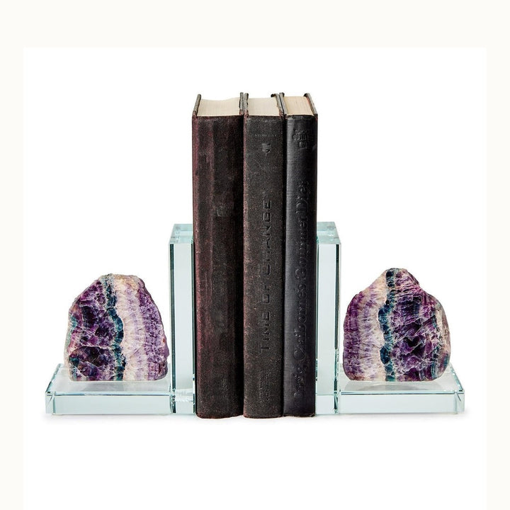 Polished glass and rainbow fluorite bookends supporting three dark vintage books against a white background featuring vibrant banded layers of purple and teal stone mounted on thick L-shaped transparent bases