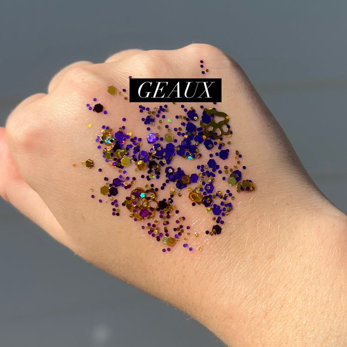 Photograph by Geaux of purple and gold chunky cosmetic glitter swatched on the back of a hand featuring shimmering hexagonal flakes, decorative gold paw print charms, and a black GEAUX text overlay