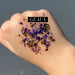 Photograph by Geaux of purple and gold chunky cosmetic glitter swatched on the back of a hand featuring shimmering hexagonal flakes, decorative gold paw print charms, and a black GEAUX text overlay