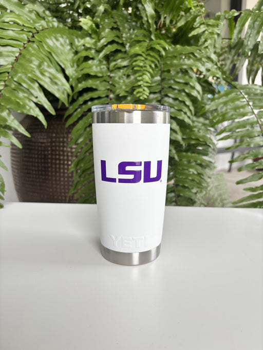 Digital photograph of a white YETI tumbler featuring a purple LSU logo and an orange magnetic slider lid sitting on a white table against a backdrop of a lush green fern and a textured brown pot