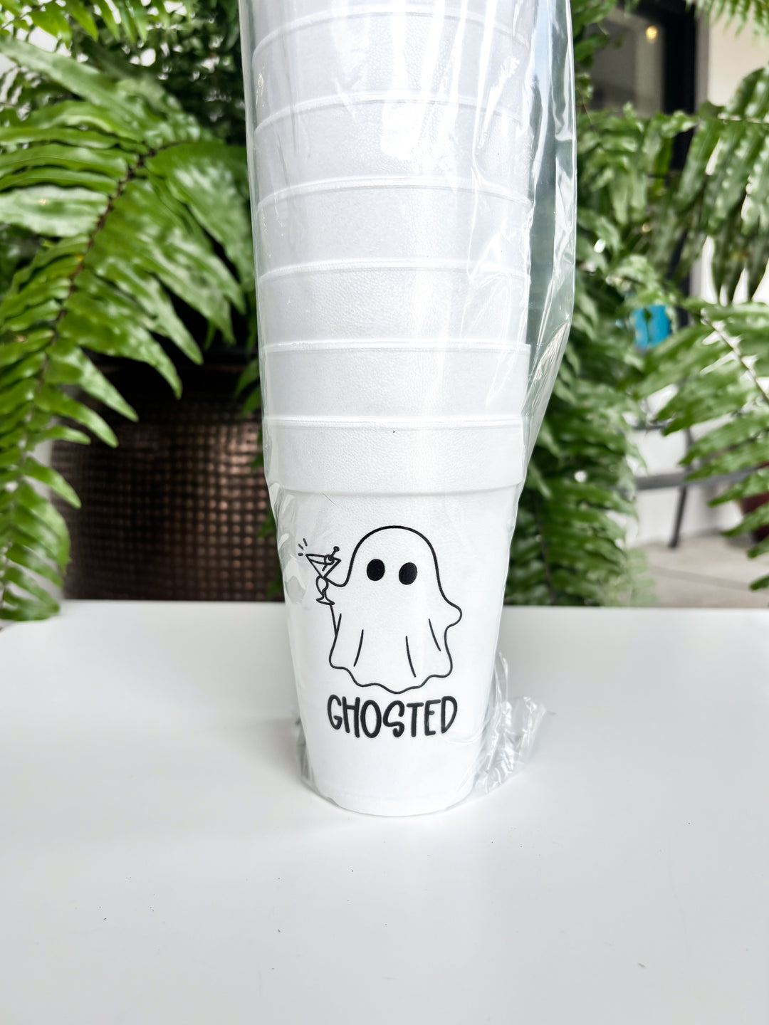 Photograph of a stack of white styrofoam cups wrapped in plastic on a white table featuring a black graphic of a ghost holding a martini glass with the word GHOSTED in front of lush green fern leaves