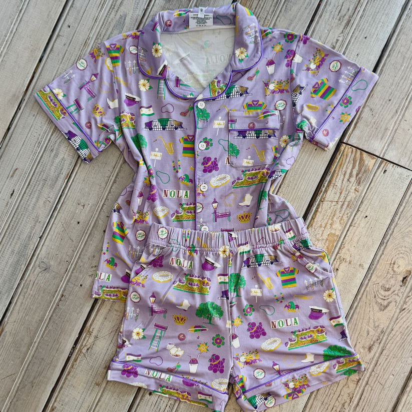 Kids' lavender short-sleeve pajama top and shorts printed with NOLA and Mardi Gras icons, laid on a wood deck