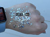Digital photograph of Silver Bells chunky iridescent glitter swatched on the back of a hand wearing a light blue Apple Watch featuring shimmering hexagonal flakes and vibrant blue reflections