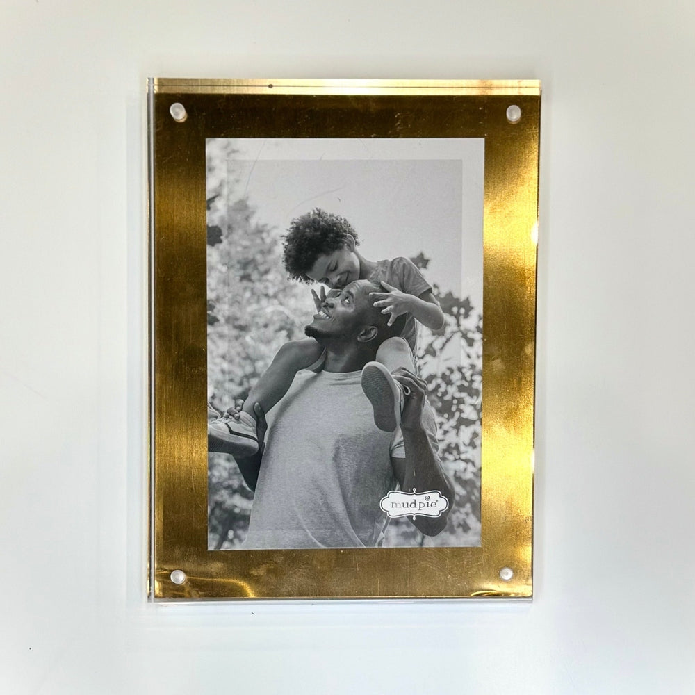 Black and white photograph by Mud Pie showing a joyful father giving his son a shoulder ride in a park, presented in a gold-bordered acrylic frame with four silver standoff screws and a leafy outdoor background