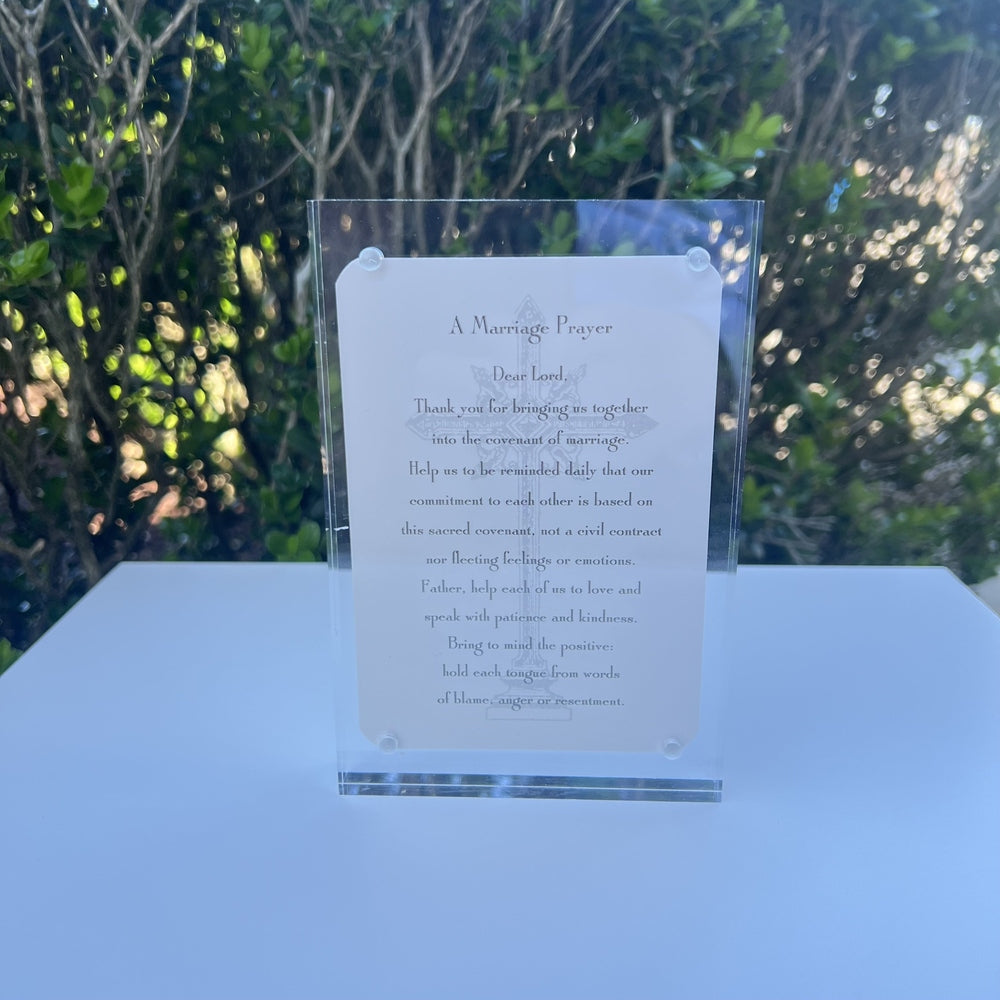 Printed marriage prayer card in a clear acrylic frame sits on a white table outdoors featuring a faint central cross illustration, silver corner fasteners, and a backdrop of sun-dappled green leaves