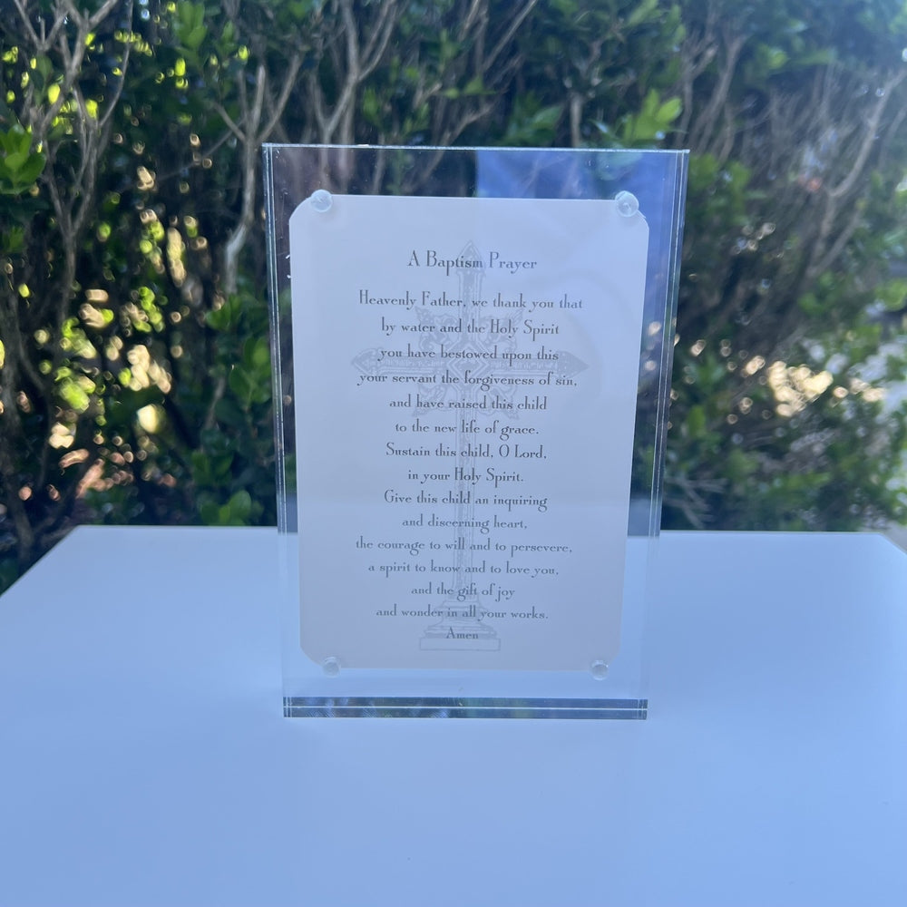 Clear acrylic block frame housing a printed baptism prayer card with a faint Celtic cross illustration is positioned on a white surface featuring four silver pins and a lush green garden background