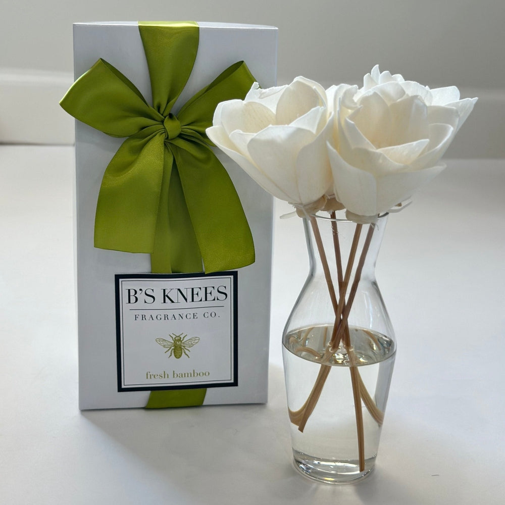 Product photography by B's Knees Fragrance Co of a fresh bamboo fragrance diffuser set on a white background featuring white sola wood flowers in a clear glass vase next to a white gift box with a lime green satin bow and a gold bee logo