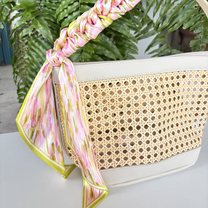Woven handbag with a floral scarf draped over it, set against a green leafy background.