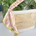 Woven handbag with a floral scarf draped over it, set against a green leafy background.