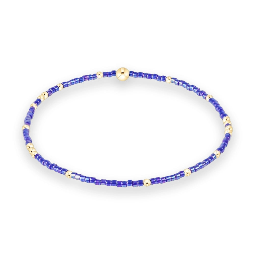 Beaded jewelry stretch bracelet centered on a white background featuring iridescent violet-blue cylindrical beads accented by polished gold-tone spacers and a large central gold sphere