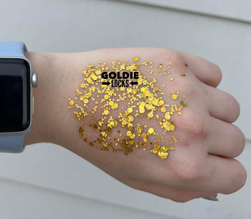 Digital photograph for Goldie Locks featuring shimmering gold hexagonal glitter of varying sizes on the back of a hand wearing an Apple Watch with a light blue band against white horizontal siding