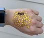 Digital photograph for Goldie Locks featuring shimmering gold hexagonal glitter of varying sizes on the back of a hand wearing an Apple Watch with a light blue band against white horizontal siding