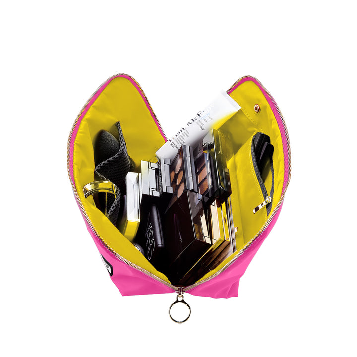 Studio photograph of a Trish McEvoy makeup bag open and filled with cosmetics on a white background, highlighting its hot pink shell, vibrant yellow lining, and circular gold zipper pull along with iconic branded products like BB balm and eyeshadow palettes