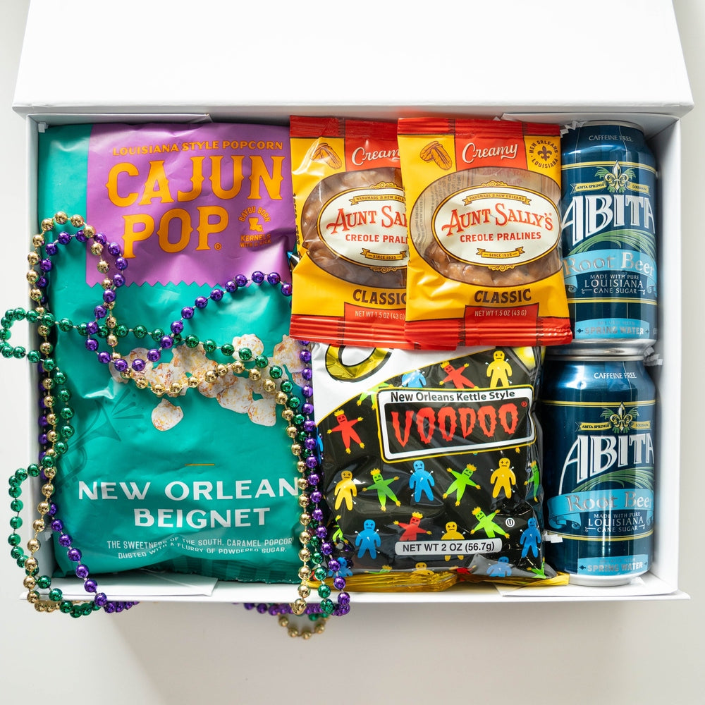 Photograph of a New Orleans-themed gift box featuring a teal bag of Cajun Pop beignet popcorn, blue cans of Abita Root Beer, and yellow packages of Aunt Sally’s Pralines draped with metallic purple, green, and gold Mardi Gras beads