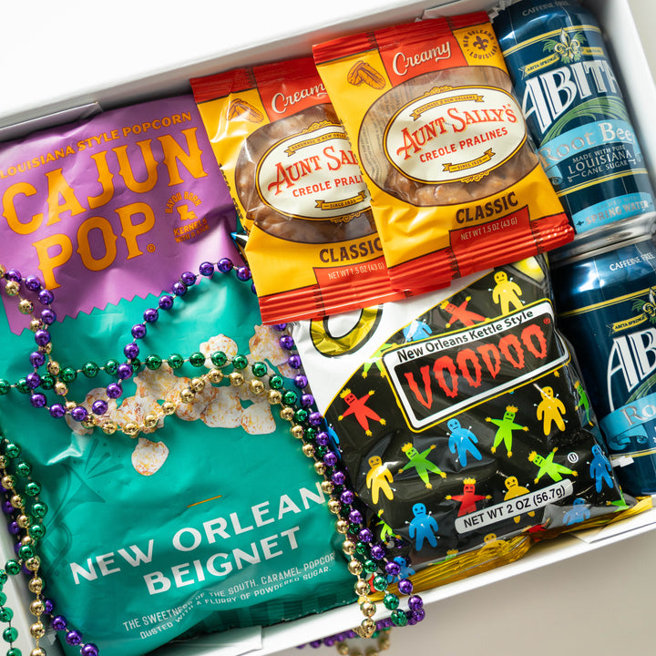 Photograph of a New Orleans snack gift box featuring Aunt Sally’s Pralines, Zapp’s Voodoo chips, and Abita Root Beer cans accented by shimmering purple, green, and gold Mardi Gras beads draped over a vibrant teal bag of beignet-flavored popcorn
