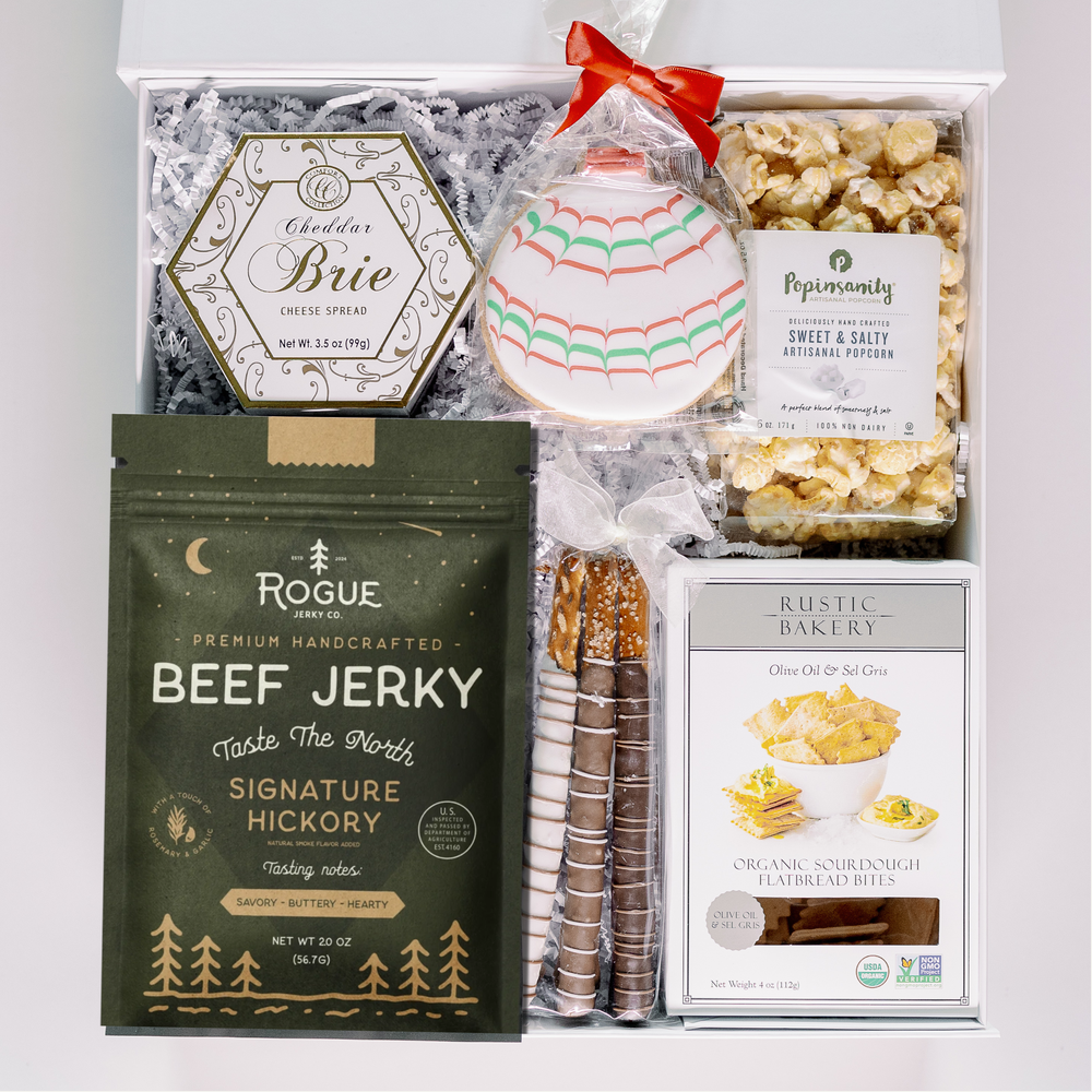 Photograph of a curated gourmet snack box featuring Rogue Jerky Co and Rustic Bakery products alongside a festive red-bowed icing cookie and chocolate-covered pretzels on white shredded paper