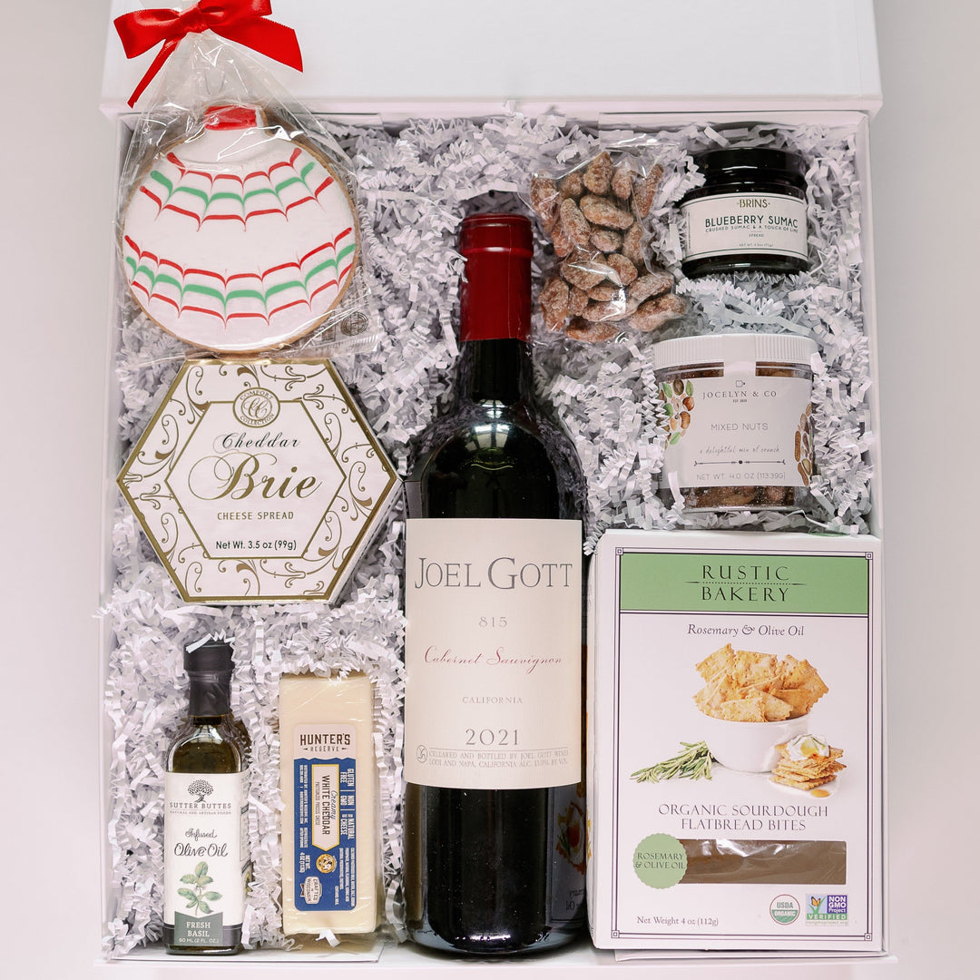 Photograph of a gourmet holiday gift box featuring a central bottle of 2021 Joel Gott Cabernet Sauvignon surrounded by a festive iced ornament cookie with a red bow, a hexagonal container of Cheddar Brie cheese spread, and a package of Rustic Bakery rosemary flatbread bites on white crinkle paper