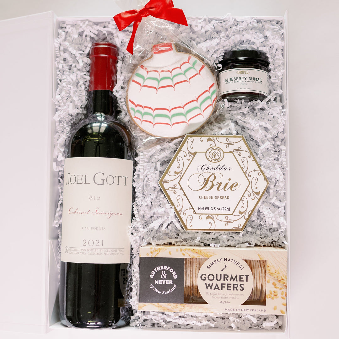 Photograph of a gourmet wine and cheese gift box featuring a bottle of Joel Gott Cabernet Sauvignon, a hexagonal Cheddar Brie container, and a decorative holiday cookie with red and green scalloped icing tied with a red ribbon