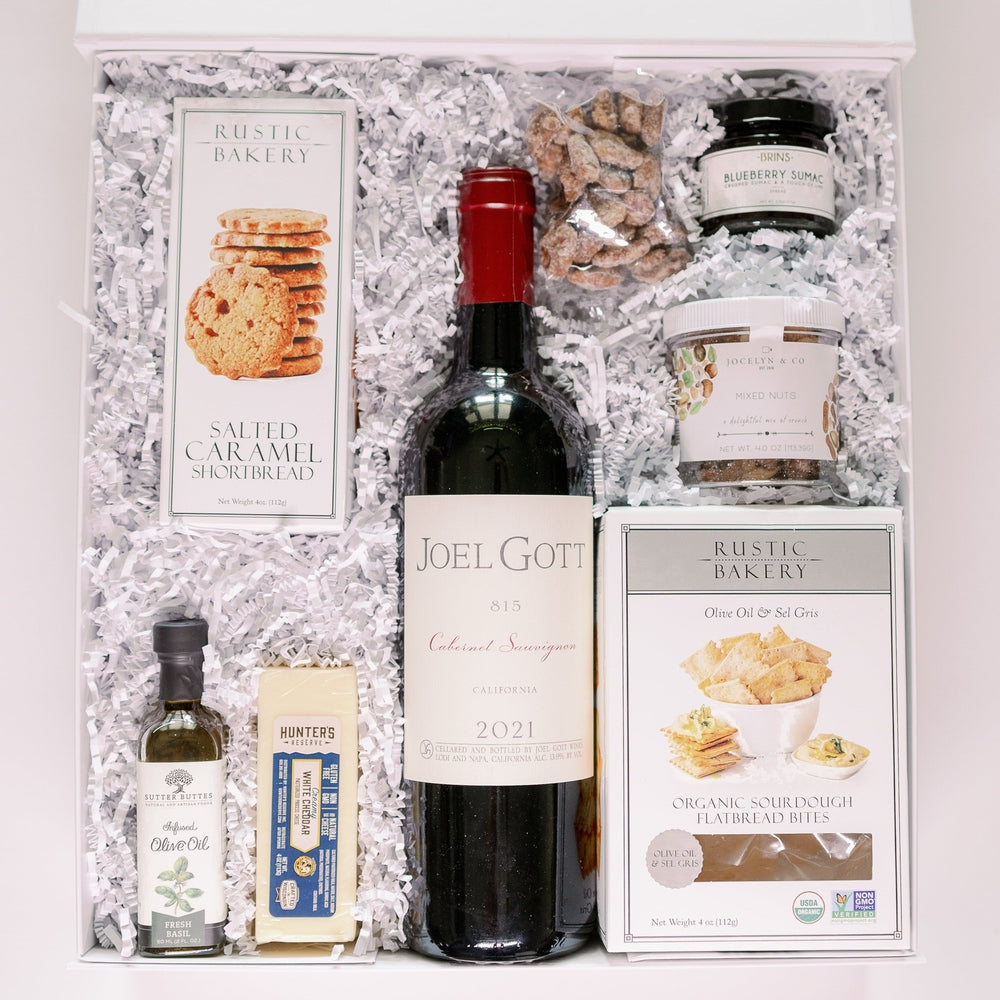 Photograph of a gourmet wine gift box featuring a central Joel Gott Cabernet Sauvignon bottle surrounded by Rustic Bakery crackers, blueberry sumac jam, and fresh basil infused olive oil on white shredded paper