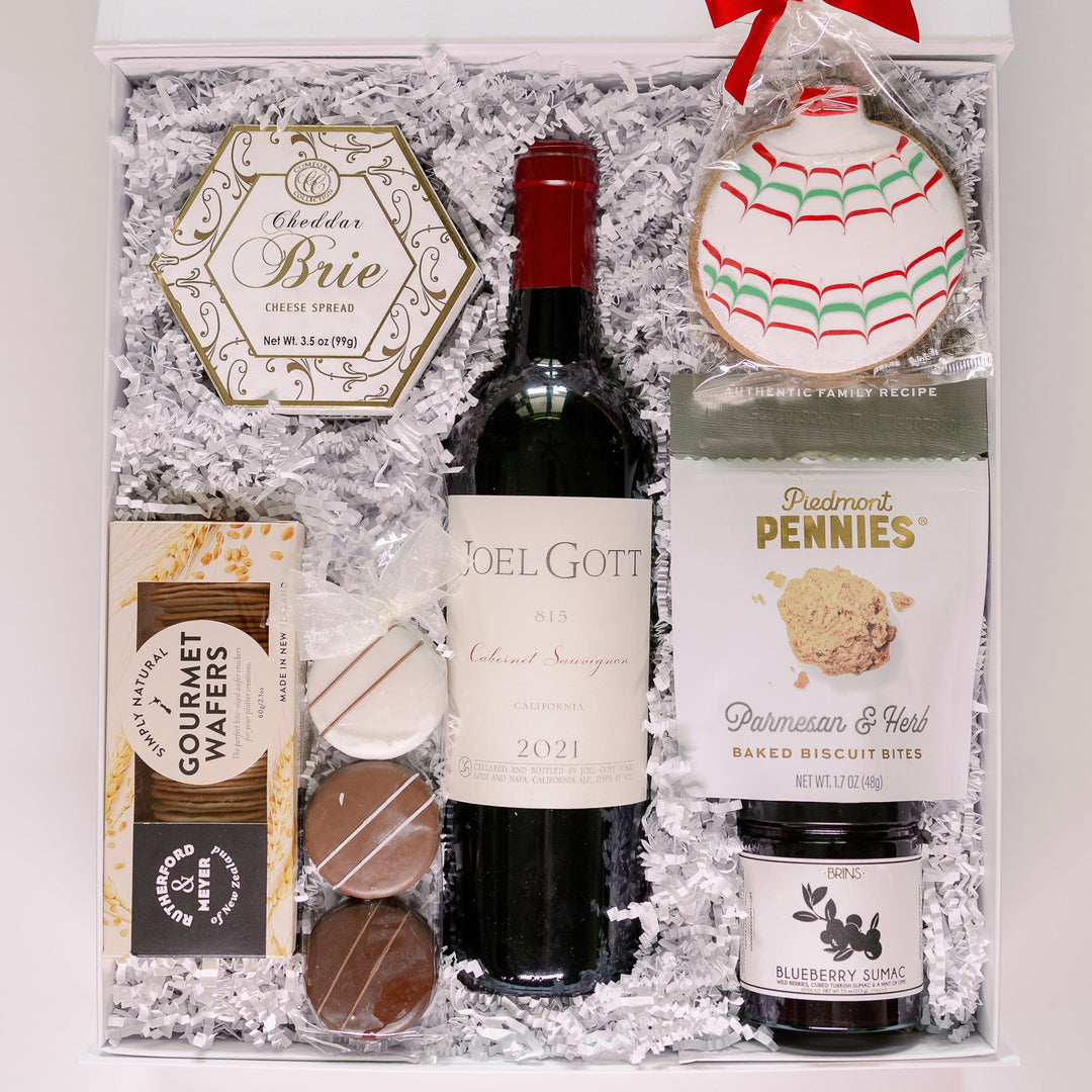 Studio photograph of a curated gourmet gift box in a white container featuring a bottle of Joel Gott 815 Cabernet Sauvignon, a festive ornament-shaped sugar cookie with red and green piping, and an elegant hexagonal tin of Cheddar Brie spread